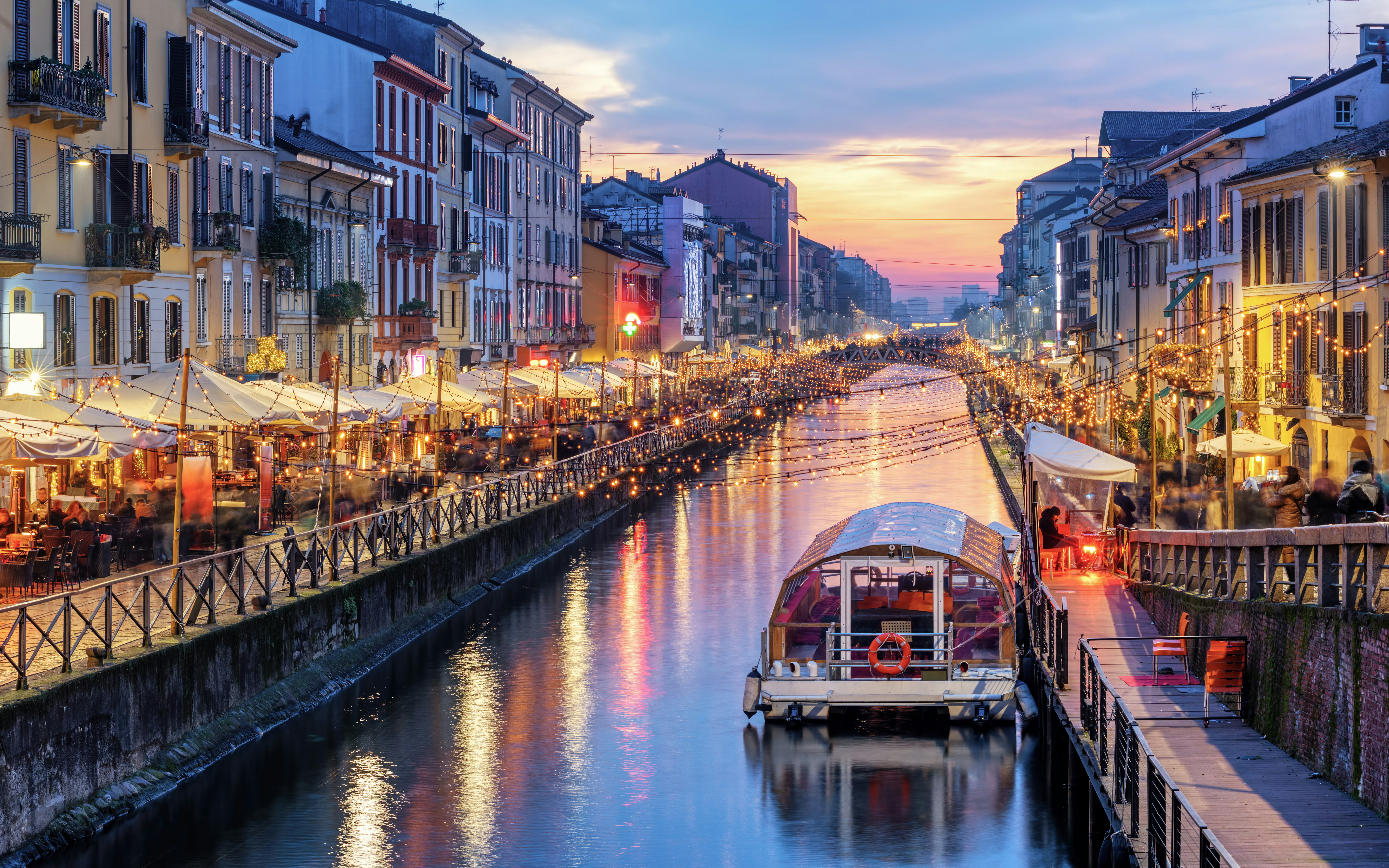 Navigli canals in Milan at sunset with lit cafes and a boat on the water.