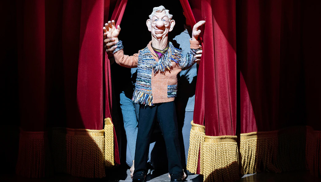 Puppet character on stage behind red curtains in "Idiots Assemble: Spitting Image the Musical.