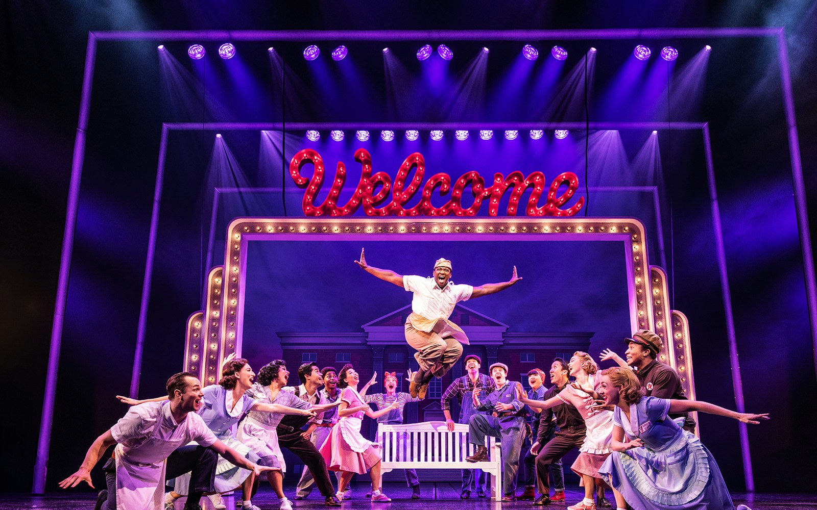 Cast performing a lively scene in Back to the Future the Musical with a "Welcome" sign.