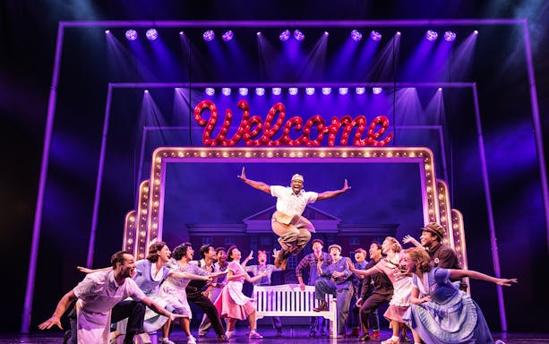 Cast performing a lively scene in Back to the Future the Musical with a "Welcome" sign.