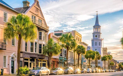 Historic Charleston street with colorful buildings and palm trees, featuring a prominent church steeple.