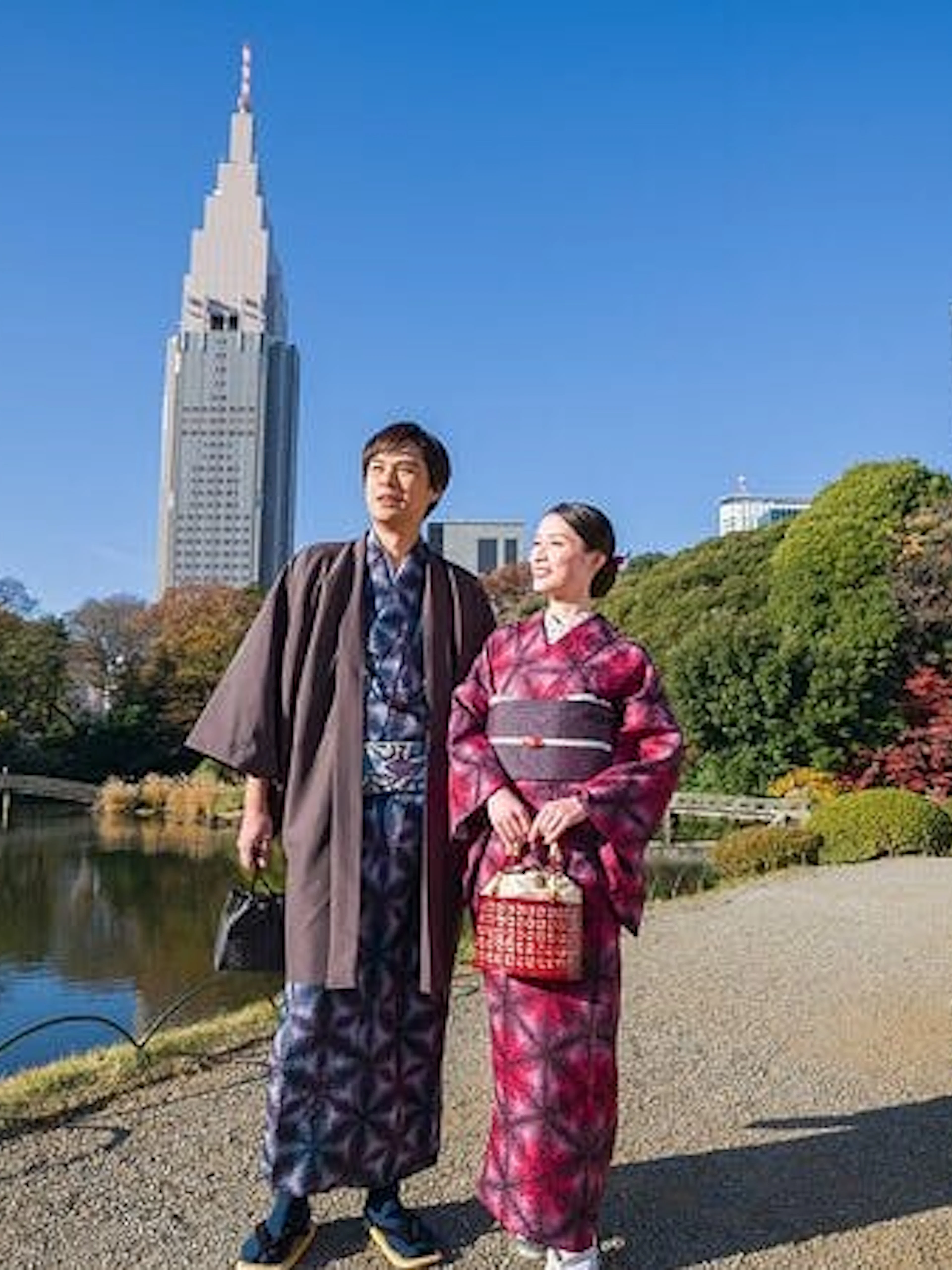 Couple in kimonos walking in Shinjuku Gyoen National Garden, Tokyo, with city skyline.