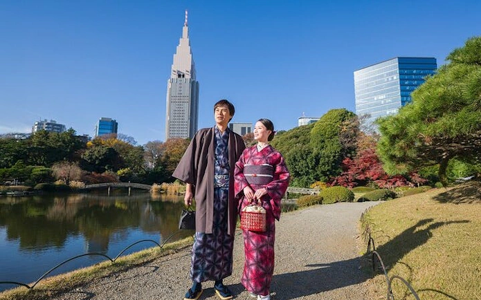 Couple in kimonos walking in Shinjuku Gyoen National Garden, Tokyo, with city skyline.
