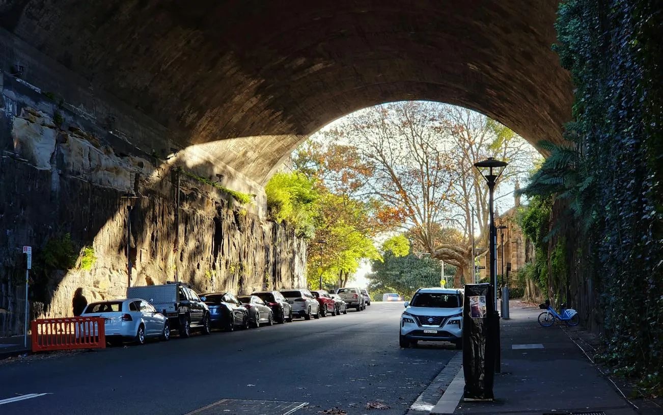 Street view under a stone archway in The Rocks, Sydney, with parked cars and trees.