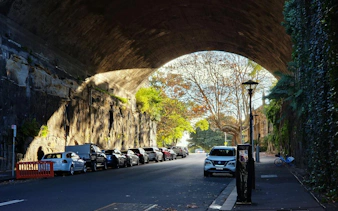 Street view under a stone archway in The Rocks, Sydney, with parked cars and trees.