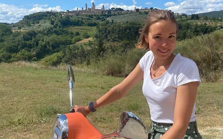 Person riding a Vespa with Tuscan hills and San Gimignano skyline in the background.