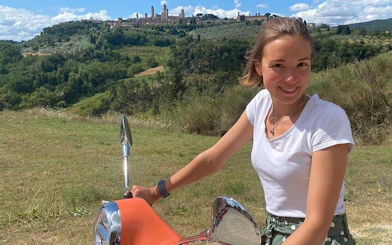 Person riding a Vespa with Tuscan hills and San Gimignano skyline in the background.