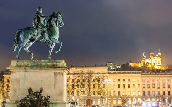 Equestrian statue in Place Bellecour, Lyon, with Basilica of Notre-Dame de Fourvière illuminated at night.