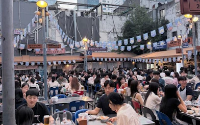 Crowded outdoor seating area in Seoul with people enjoying fried chicken and beer.