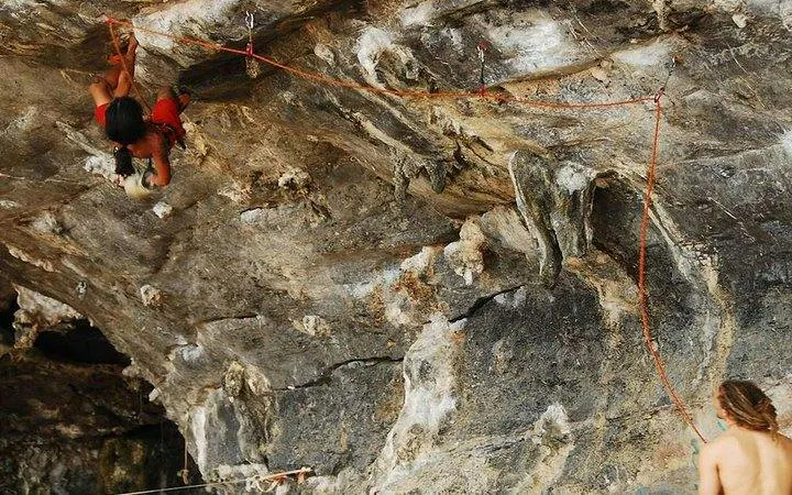 Rock climber scaling limestone cliff at Railay Beach, Krabi, with guide below.