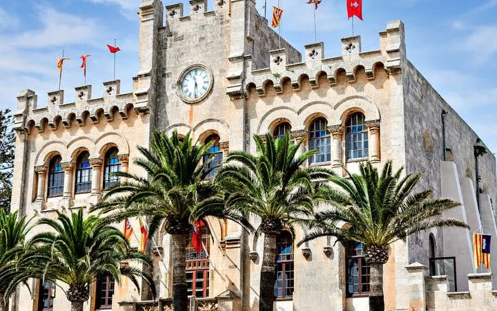 Historic building with clock tower and palm trees in Menorca, featured on self-guided walking tour app.