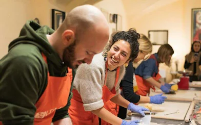 People in a Bologna cooking class making pasta, wearing aprons and gloves, smiling and interacting.