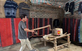 Man using traditional mill in Sapa village, surrounded by colorful textiles.