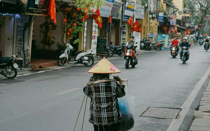 Hanoi street scene with a vendor carrying goods, motorbikes, and traditional flags.
