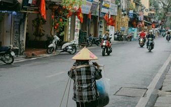 Hanoi street scene with a vendor carrying goods, motorbikes, and traditional flags.
