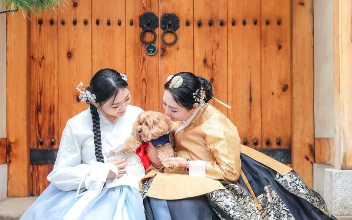 Two women in traditional hanbok with a dog at a wooden palace gate in Seoul.