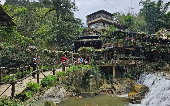 Tourists crossing a bridge near a waterfall and rustic building in Sapa village.