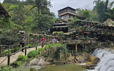 Tourists crossing a bridge near a waterfall and rustic building in Sapa village.