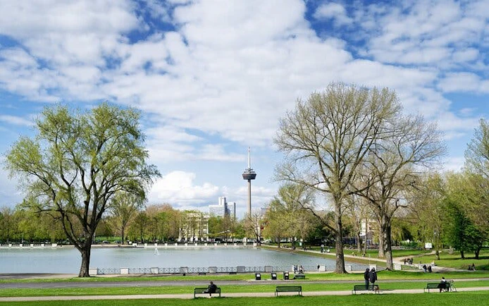 Cologne park with trees and pond, TV tower in background, part of haunted city exploration game.