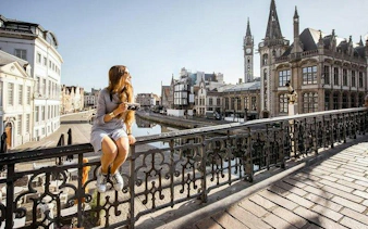 Woman sitting on a bridge railing in Bruges, with historical buildings and canal in the background.