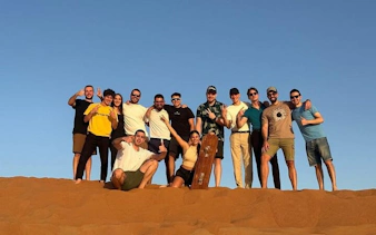Group enjoying sandboarding on a morning desert safari with dune bashing and camel ride.