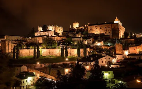 Cáceres historic cityscape illuminated at night, featuring ancient stone buildings and towers.