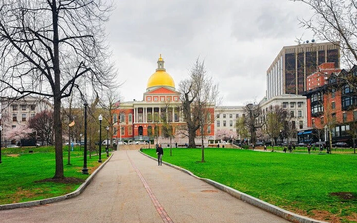 Massachusetts State House with golden dome in Boston, surrounded by park and city buildings.
