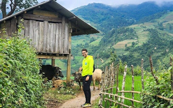 Man walking on a path beside a wooden hut and cows in Sapa, Vietnam, with terraced hills in the background.