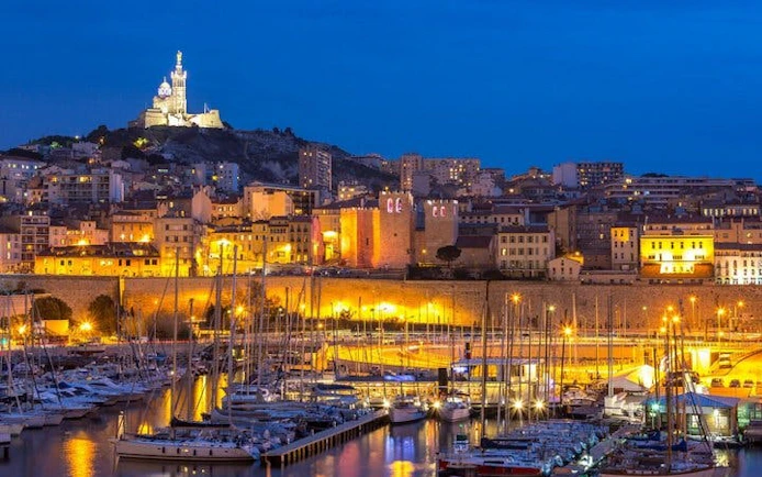 Marseille harbor at night with Notre-Dame de la Garde basilica illuminated in the background.