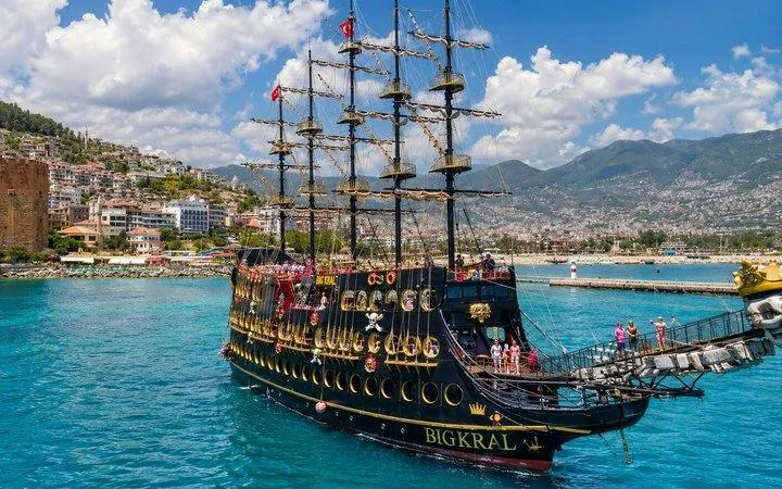 Pirate ship sailing near Alanya coast with tourists on deck, mountains in background.