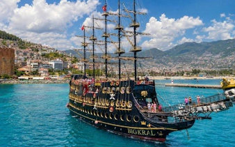 Pirate ship sailing near Alanya coast with tourists on deck, mountains in background.