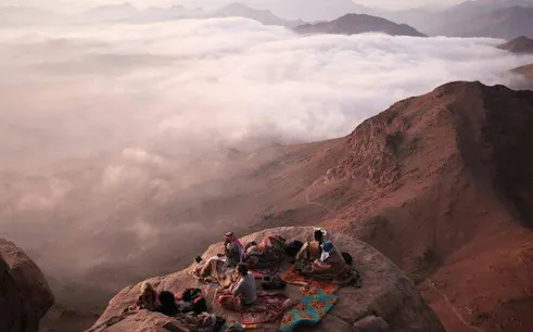 People resting on Mount Moses summit with a view of surrounding mountains and clouds, Sinai Peninsula.