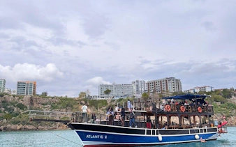 Boat tour near Antalya's Lower Duden Waterfall with passengers on deck, cityscape in background.