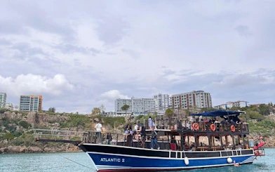 Boat tour near Antalya's Lower Duden Waterfall with passengers on deck, cityscape in background.