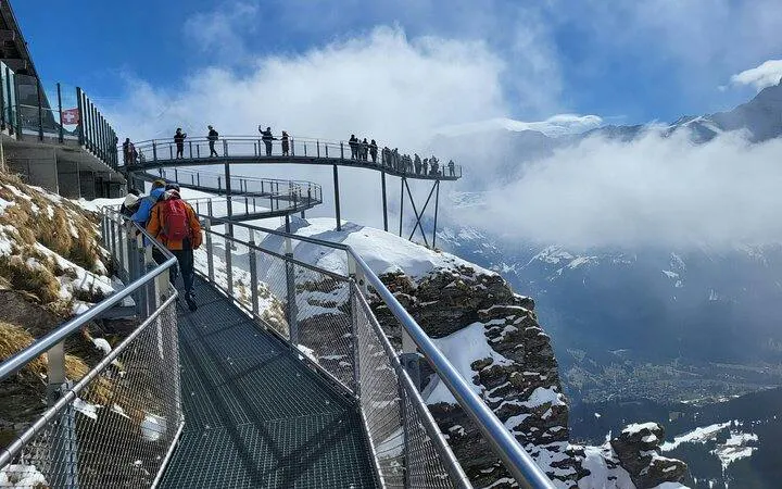 Visitors walking on a cliffside walkway at Mount Titlis, Swiss Alps, with snowy peaks and clouds.