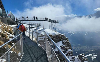 Visitors walking on a cliffside walkway at Mount Titlis, Swiss Alps, with snowy peaks and clouds.