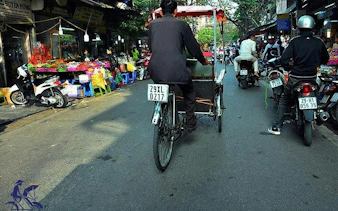 Cyclo navigating Hanoi Old Quarter street market with motorbikes and colorful stalls.