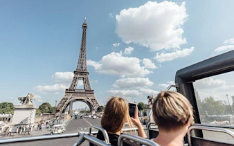 Open-top bus view of Eiffel Tower, Paris, with tourists capturing photos.