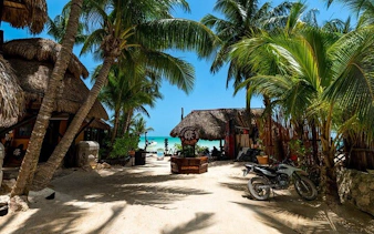 Holbox Island beach entrance with palm trees and thatched huts, leading to turquoise sea.