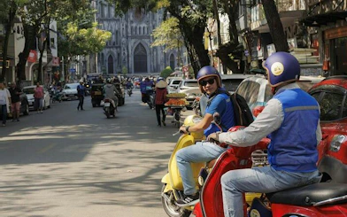 Vespa riders on a street in Hanoi with St. Joseph's Cathedral in the background.
