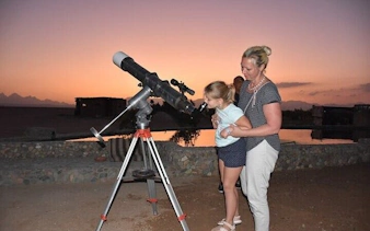 Child and adult stargazing through telescope at sunset in Sharm El Sheikh desert.