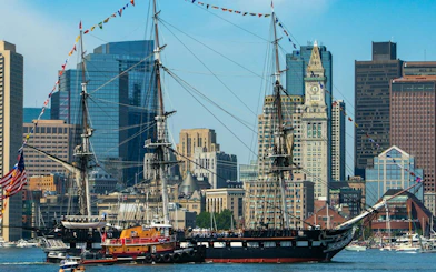 Historic ship USS Constitution in Boston Harbor with city skyline in the background.