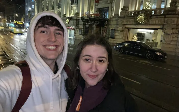 Selfie of two people on a nighttime street in Manchester, with festive lights and a tram in the background.