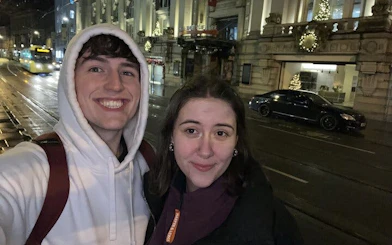 Selfie of two people on a nighttime street in Manchester, with festive lights and a tram in the background.