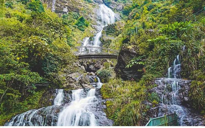 Waterfall cascading over rocks with a bridge in Sapa, Vietnam, surrounded by lush greenery.