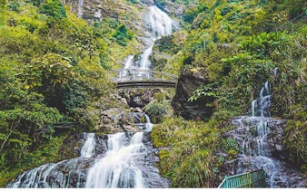 Waterfall cascading over rocks with a bridge in Sapa, Vietnam, surrounded by lush greenery.