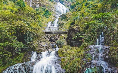 Waterfall cascading over rocks with a bridge in Sapa, Vietnam, surrounded by lush greenery.