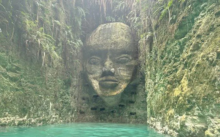 Stone face sculpture in a hidden cenote, Punta Cana, surrounded by lush vegetation.