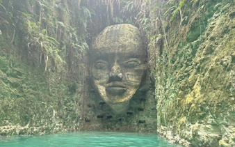 Stone face sculpture in a hidden cenote, Punta Cana, surrounded by lush vegetation.
