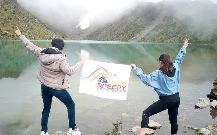Two people holding a tour operator sign at Humantay Lagoon, Cusco, with misty mountains in the background.